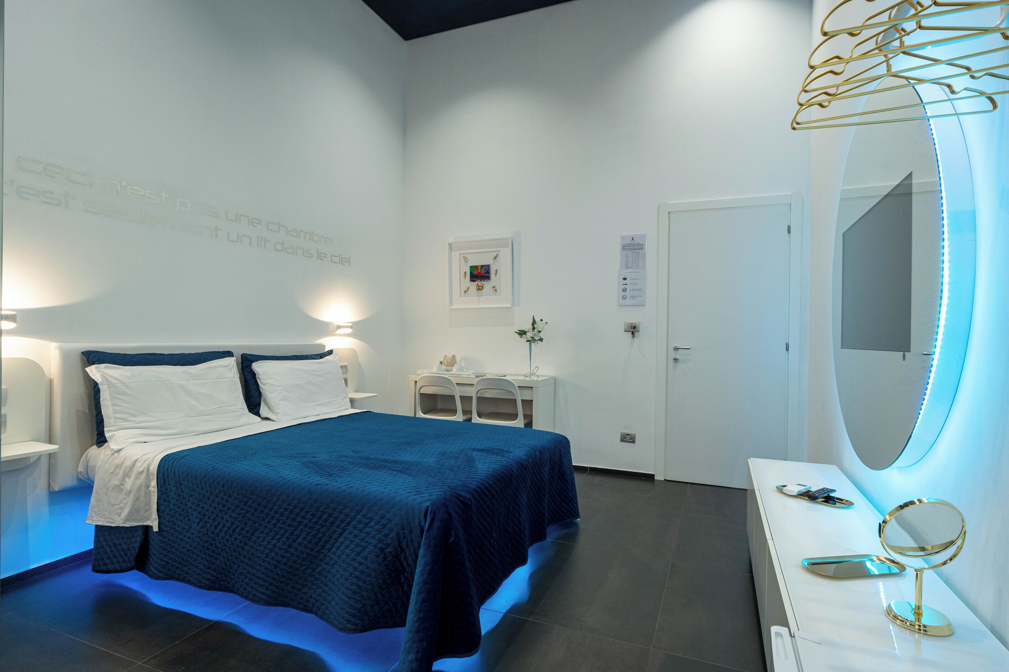 A modern bedroom with a bed, bedside tables, a desk, a mirror, and a wall-mounted light fixture.