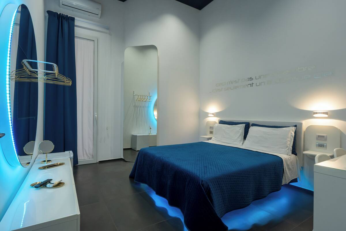 Moderni hotellihuone, jossa on suuri sänky, yöpöydät, peili ja seinään kiinnitetty valaisin.