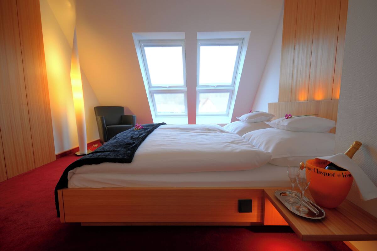 A hotel room with a large bed, a bedside table with a champagne bottle, and a window with a view.