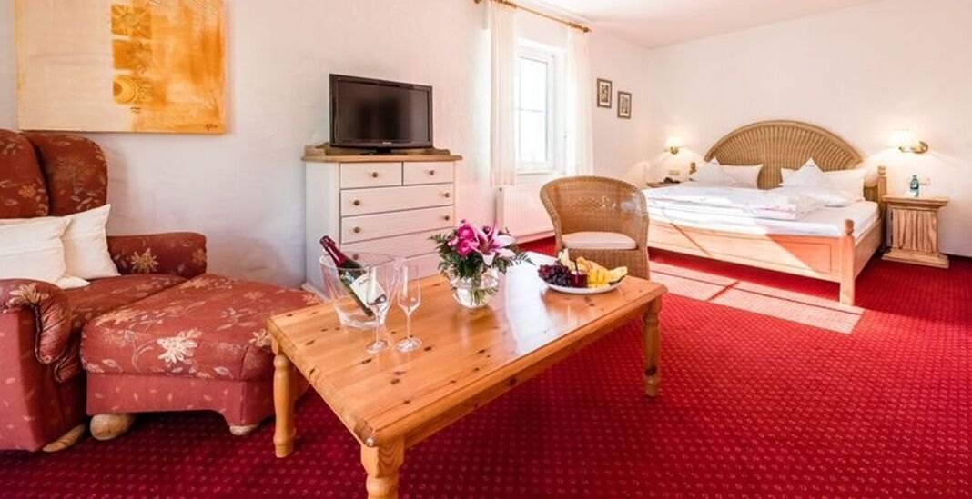 Habitación de hotel con una mesa de comedor de madera, copas y un plato de fruta, un sofá, un televisor en una cómoda y una cama con cabecera.