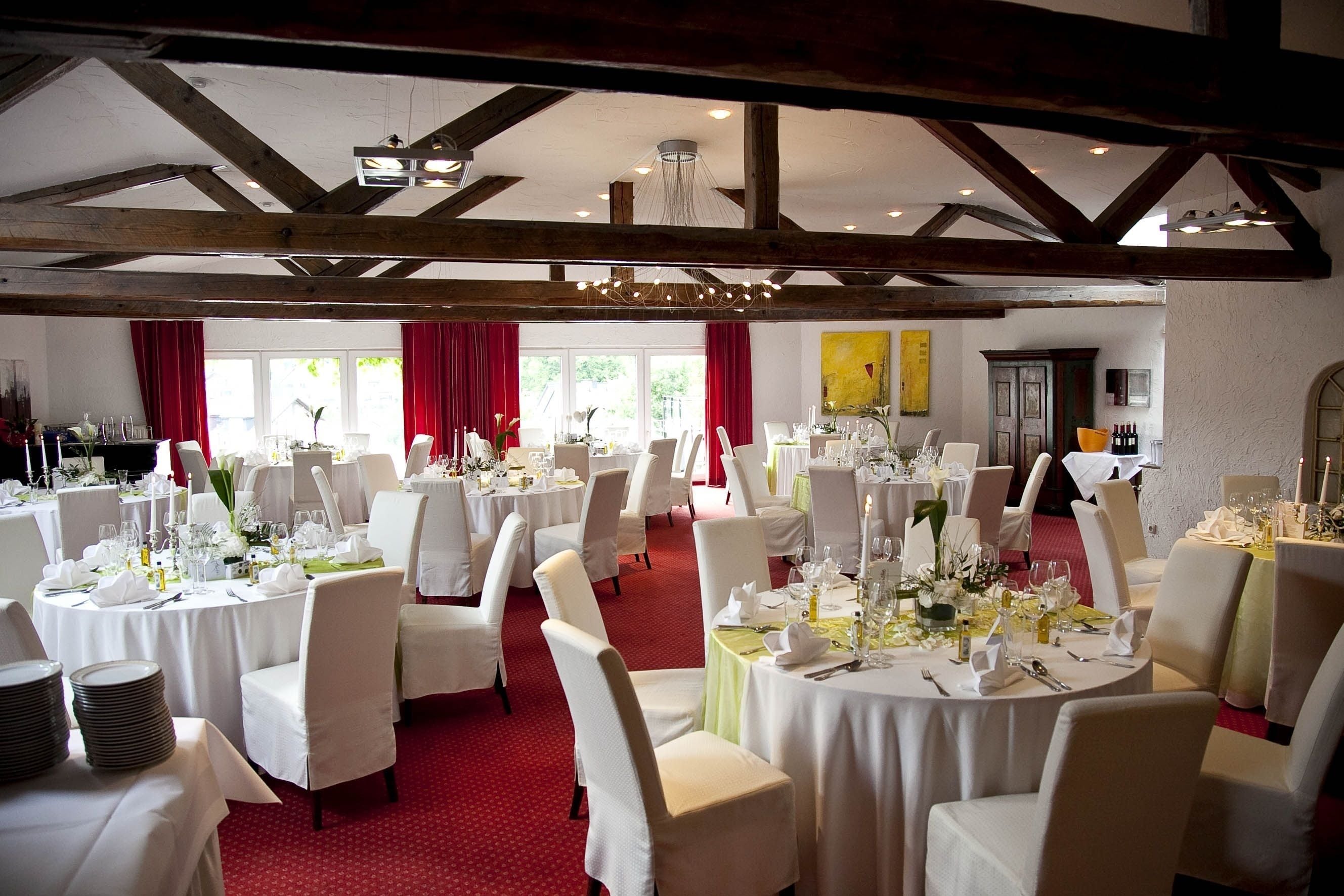 Une salle de banquet avec des tables rondes dressées pour le repas, des chaises blanches et des rideaux rouges.