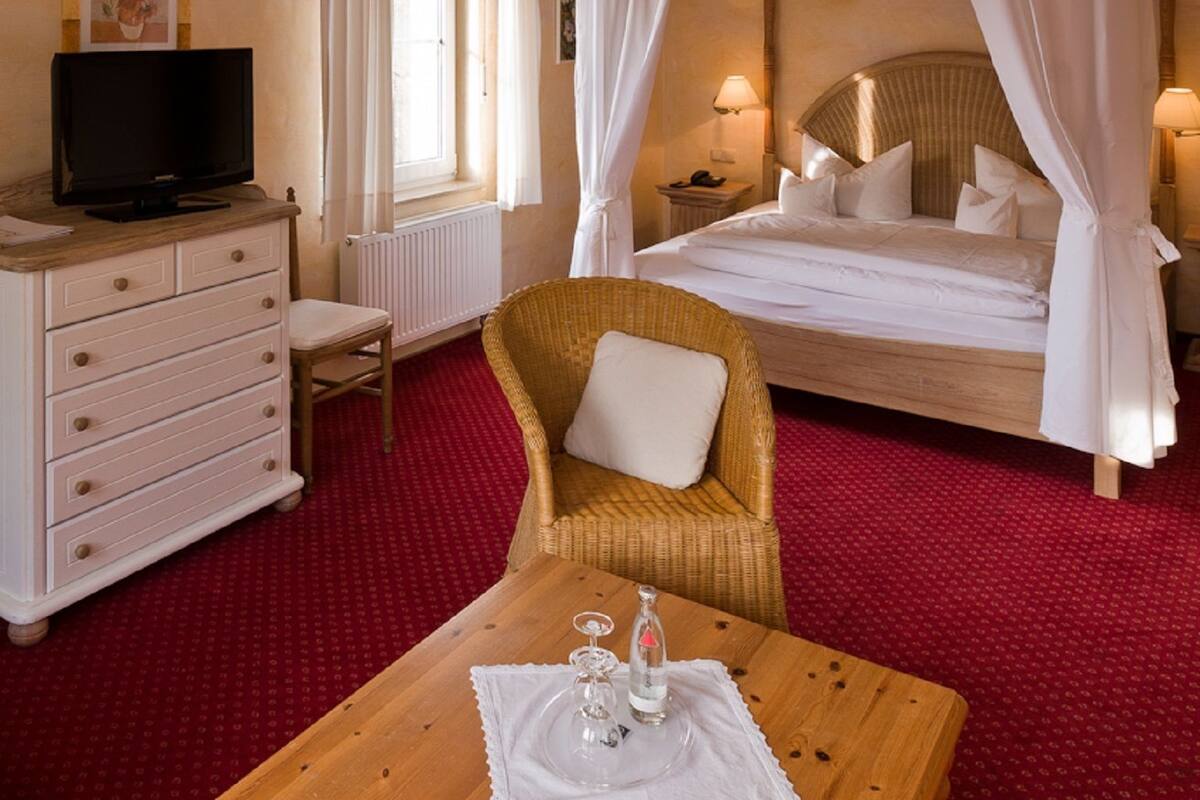 A hotel room with a bed, a television, a wicker chair, a wooden table with a glass, and a red carpet.
