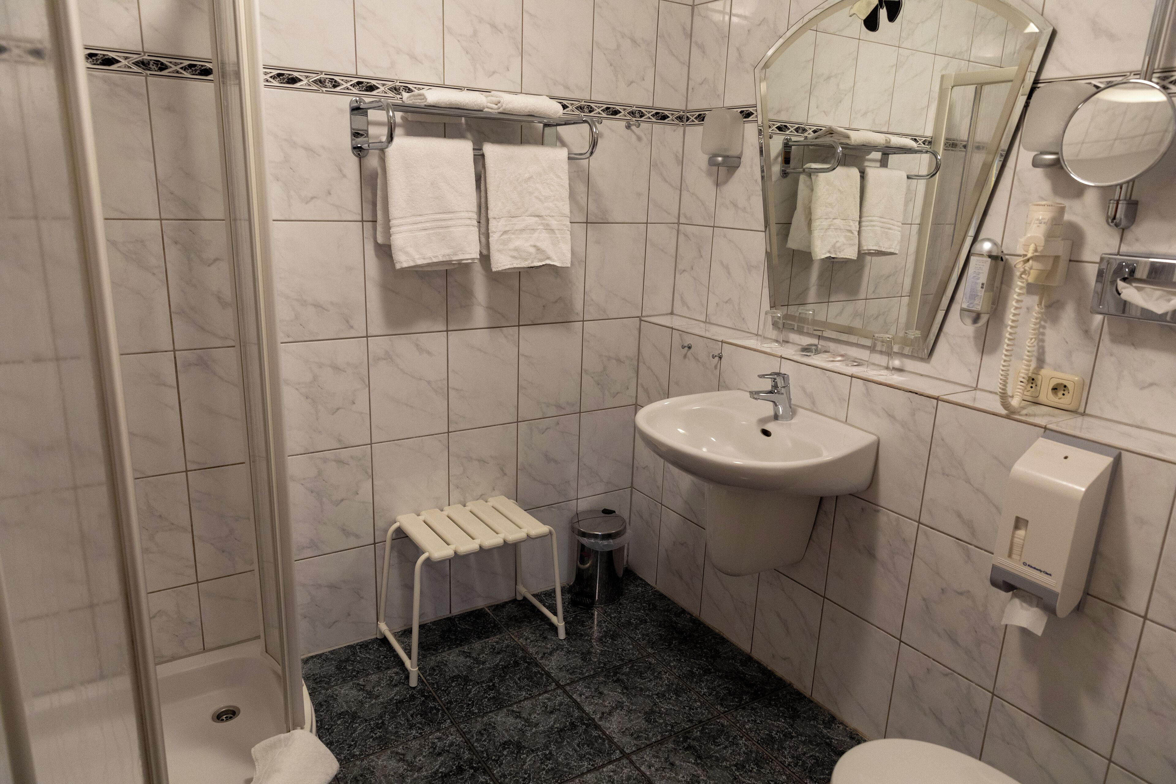 A bathroom with a shower, sink, and toilet. There are towels hanging on a rack, a trash can, and a soap dispenser.