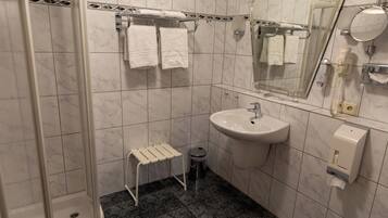 A bathroom with a shower, sink, and toilet. There are towels hanging on a rack, a trash can, and a soap dispenser.
