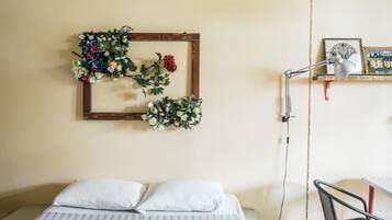 A bedroom with a bed, pillows, a desk with a chair, a desk lamp, a framed picture, and a wall-mounted floral arrangement.