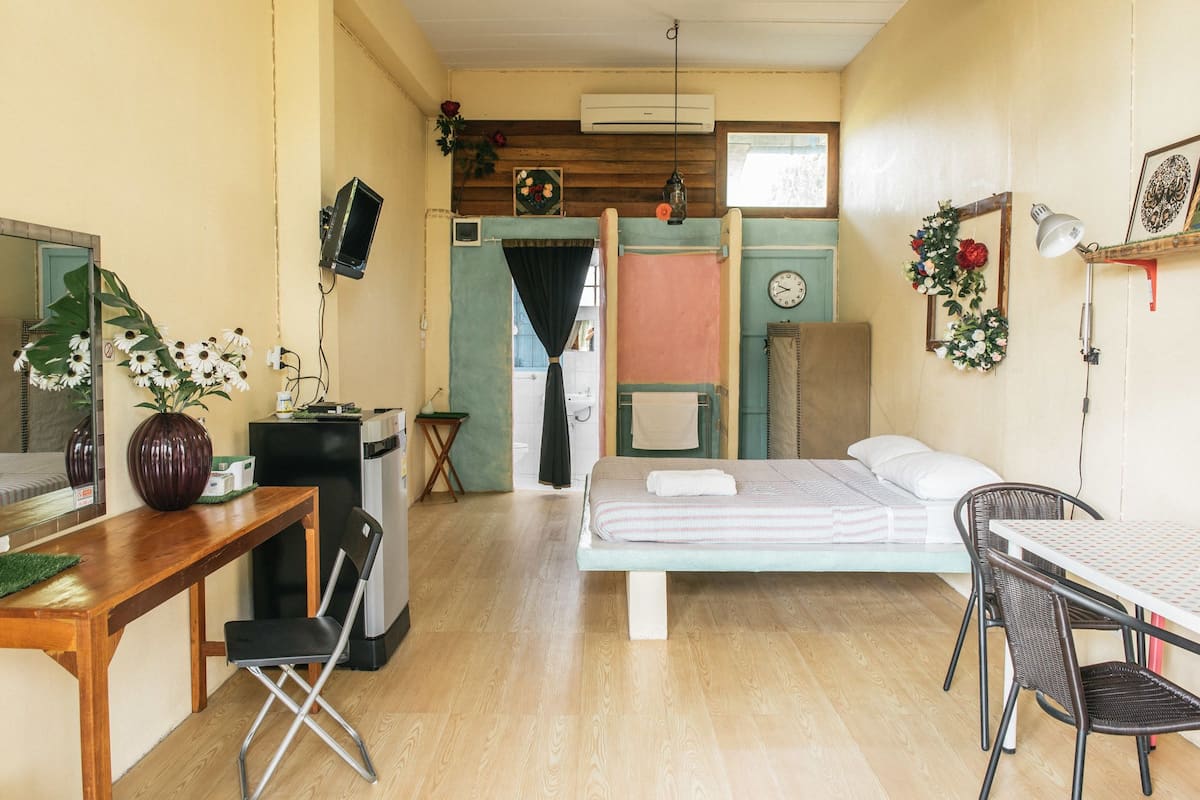 Quarto compacto com cama, escrivaninha, cadeira, mesa pequena e espelho.