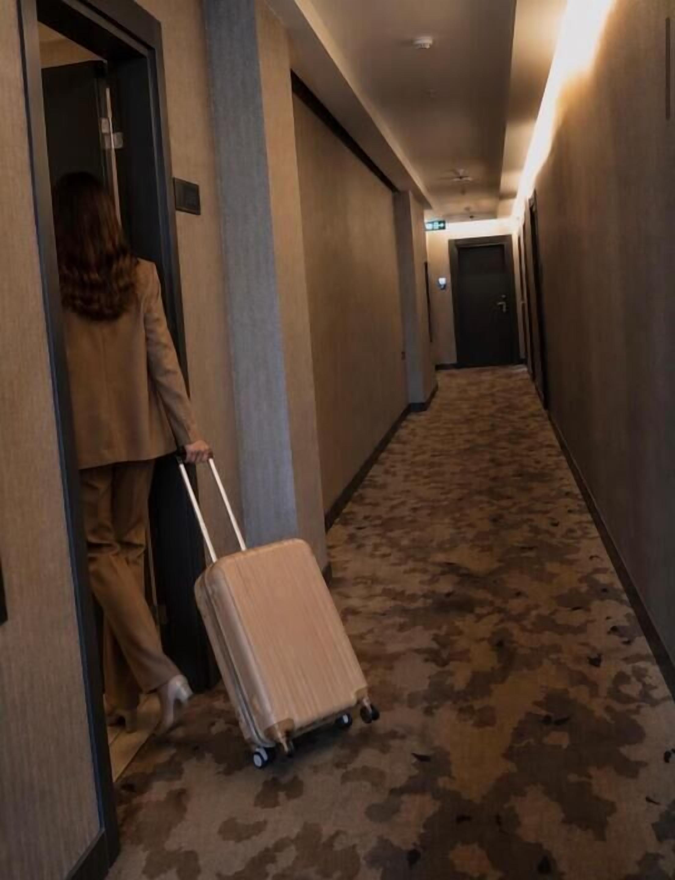 Un couloir avec un sol recouvert de moquette, une personne tenant une valise et des portes sur le côté droit.