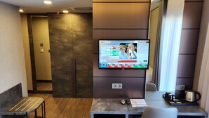 Habitación de hotel con televisión instalada en la pared mostrando un noticiero, un refrigerador pequeño con bebidas, un escritorio con una silla y una ventana con cortinas.