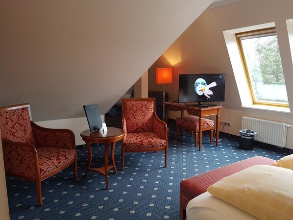 A hotel room with a bed, two armchairs, a small table, a television, and a window with a view of trees. - Dünenhotel am Meer (Rostock)
