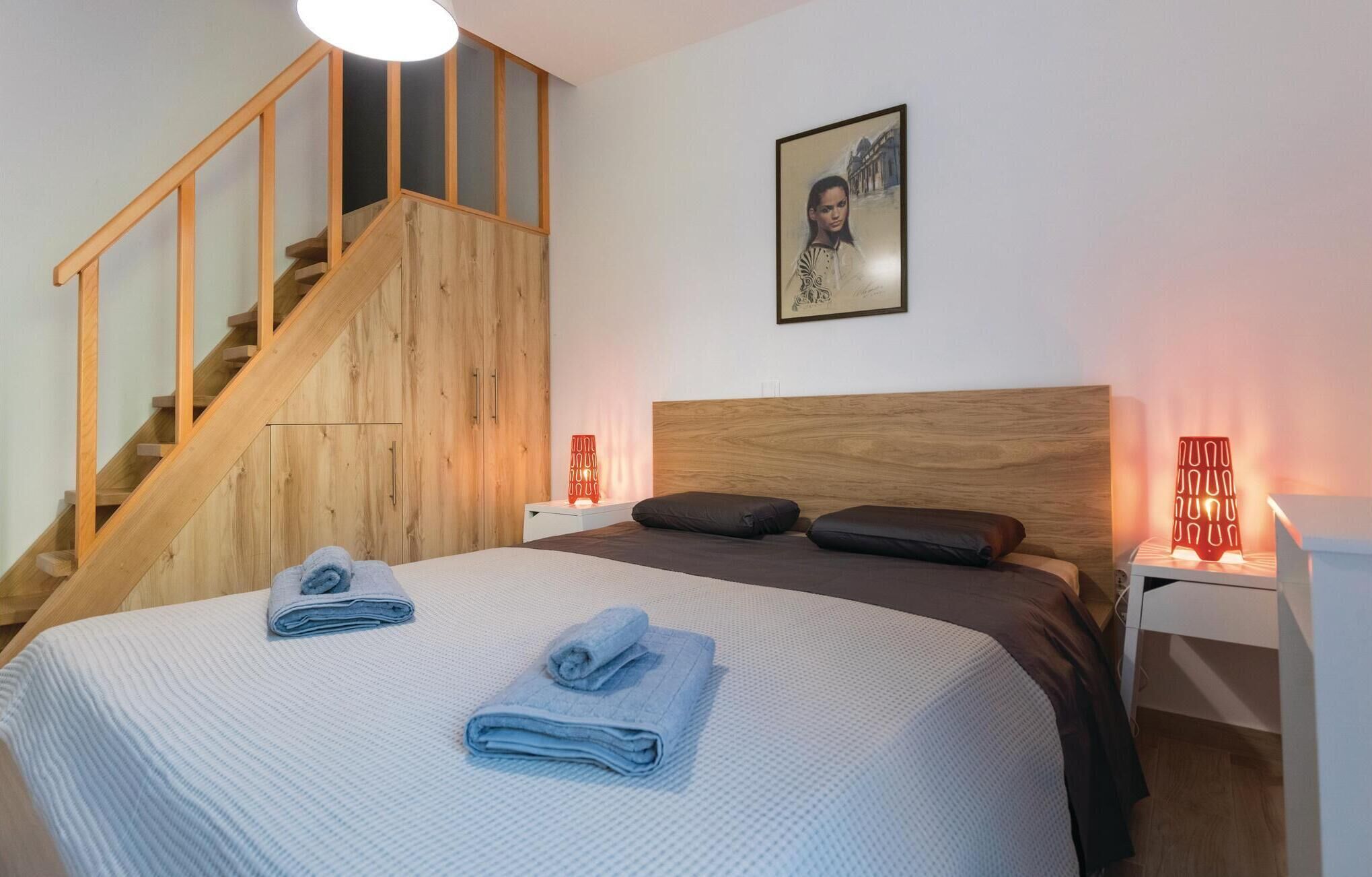 Une chambre avec un escalier en bois, un lit avec une tête de lit, deux tables de chevet avec des lampes rouges et un tableau encadré au mur.