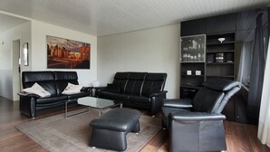 A modern living room with a black leather sofa set, a glass coffee table, and a black leather ottoman. There is a cityscape painting on the wall and a window with curtains. - Holiday house in Duhnen with a large garden and lots of space (Cuxhaven)