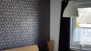 A bedroom with a large bed, a patterned wallpaper, a window with curtains, a bedside lamp, and a radiator. - Holiday house in Duhnen with a large garden and lots of space (Cuxhaven)