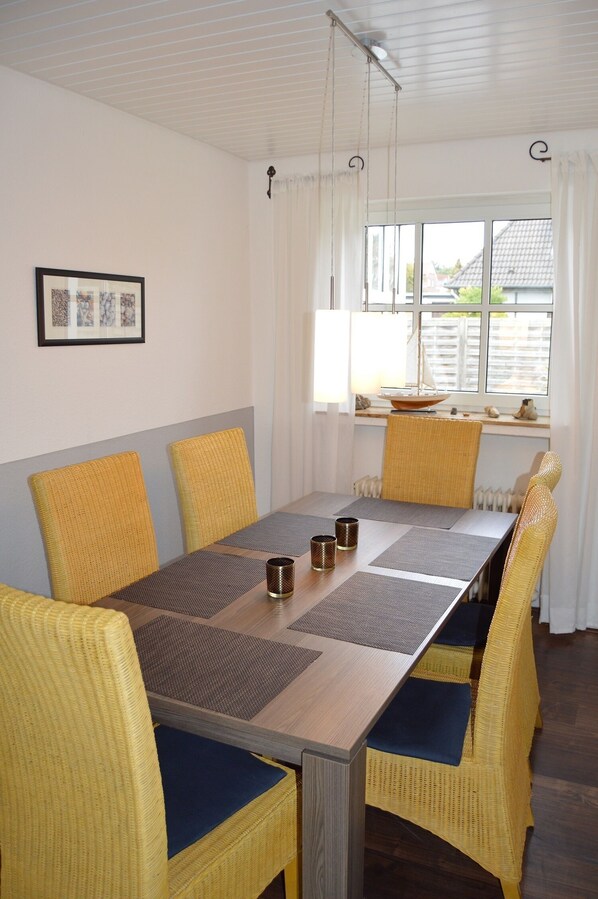 Uma área de jantar com uma mesa de madeira comprida, cadeiras de vime amarelo com almofadas azuis e um lustre pendurado.