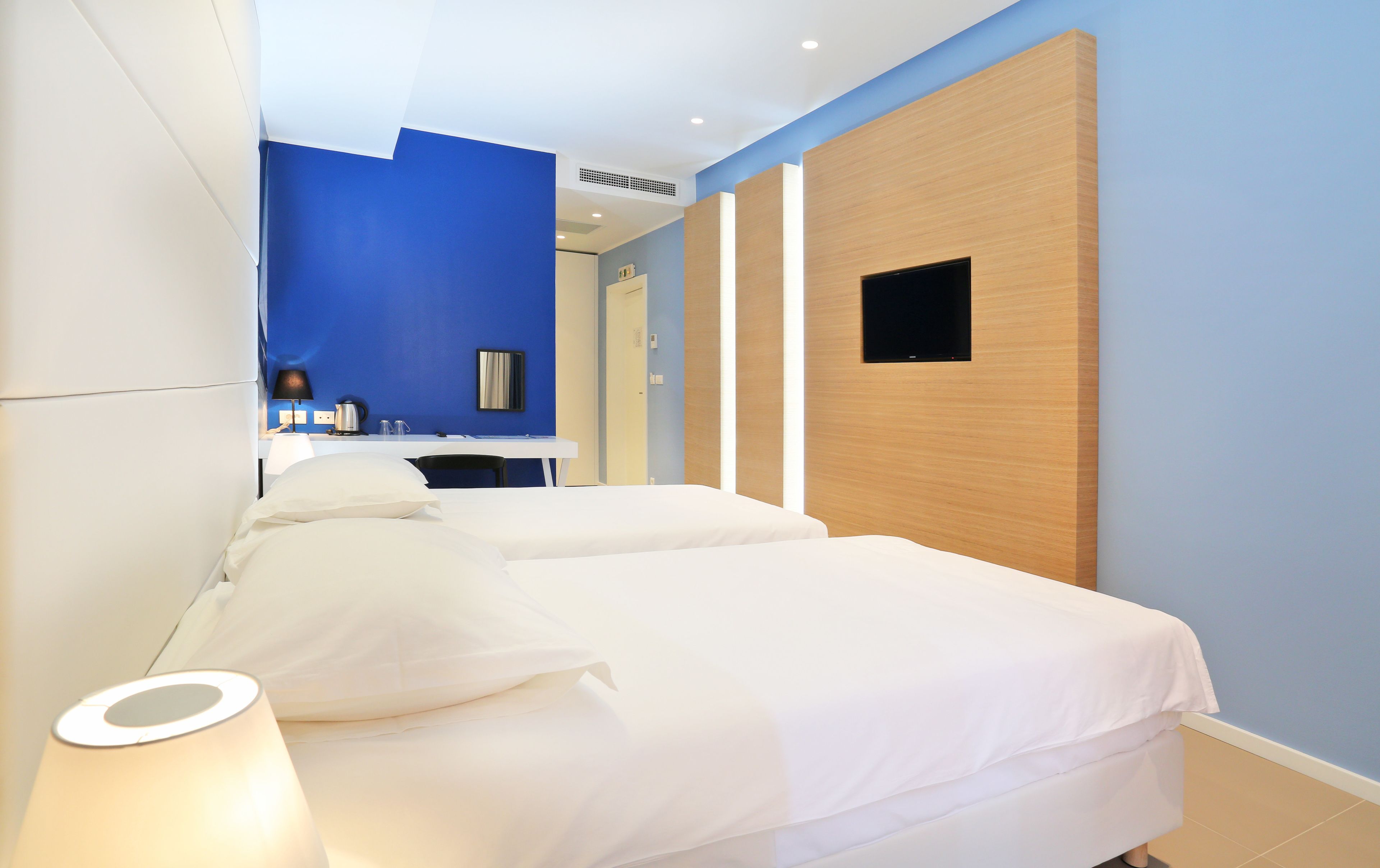 A hotel room with two beds, a desk, a chair, a flat-screen TV, and a small mirror on the wall.
