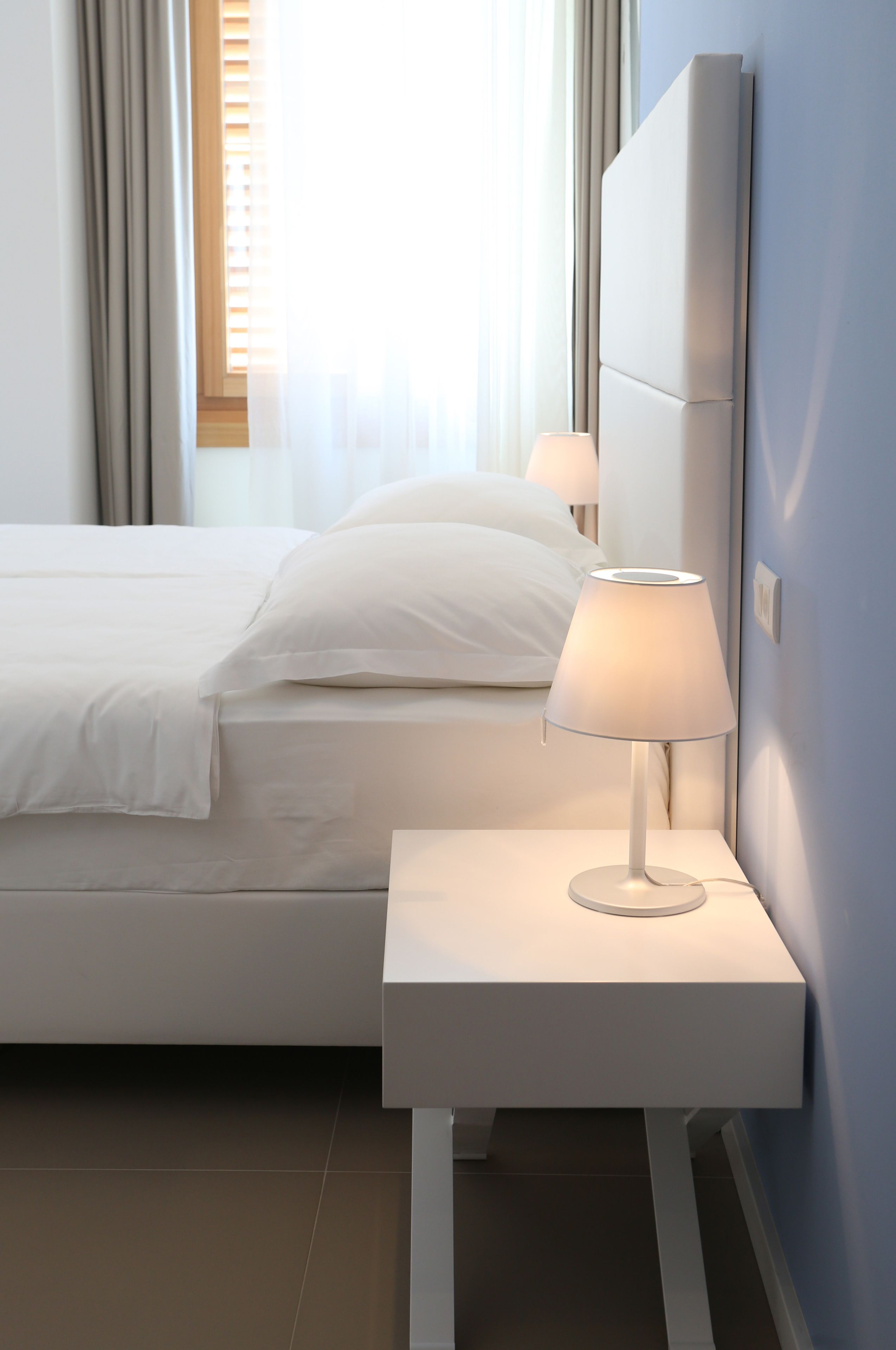 A neatly made bed with white linens and pillows, a bedside table with a lamp, and a large mirror on the wall.
