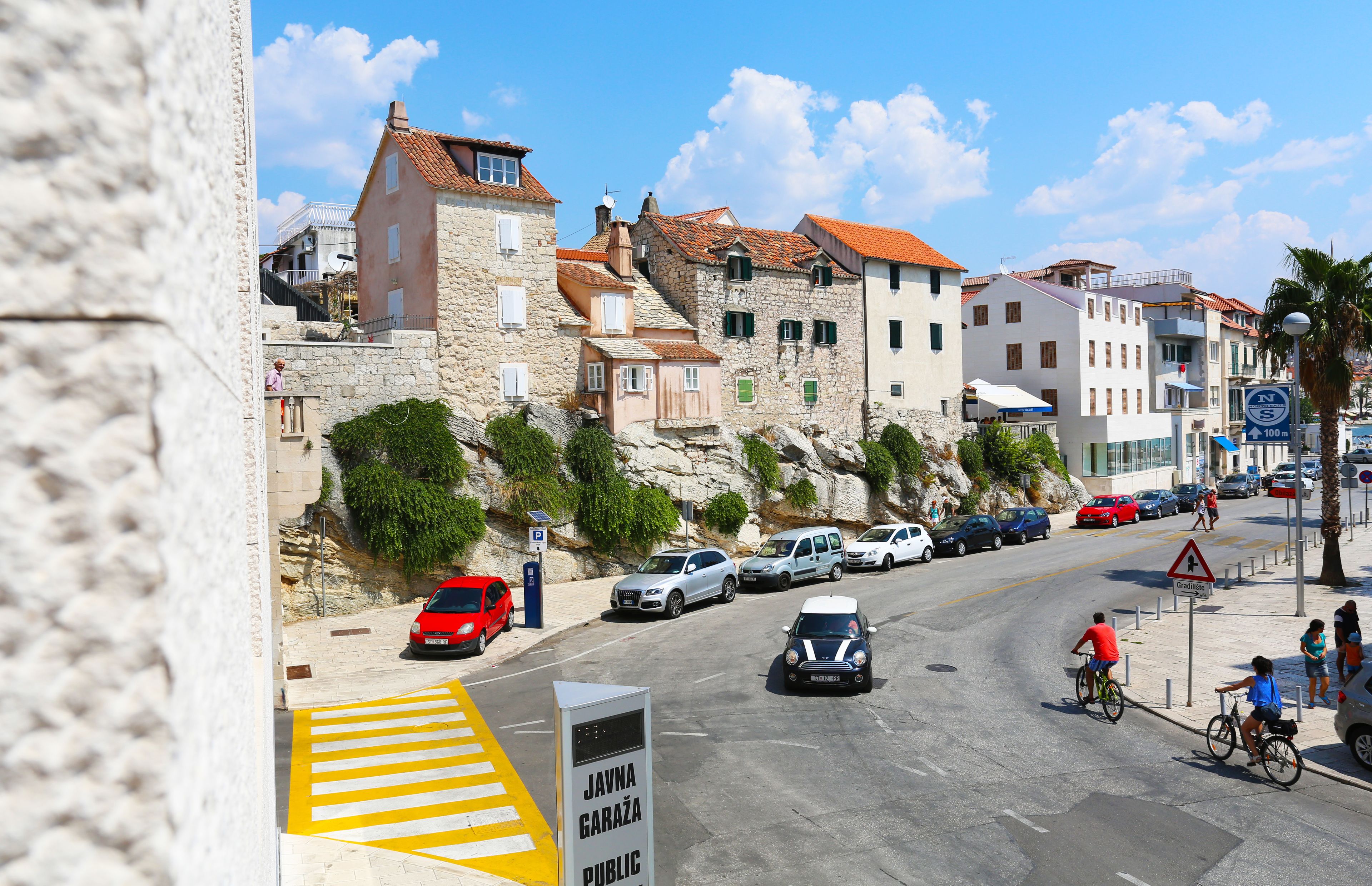 A street with cars and cyclists, stone buildings, and a sign for Javno Garaža Public.
