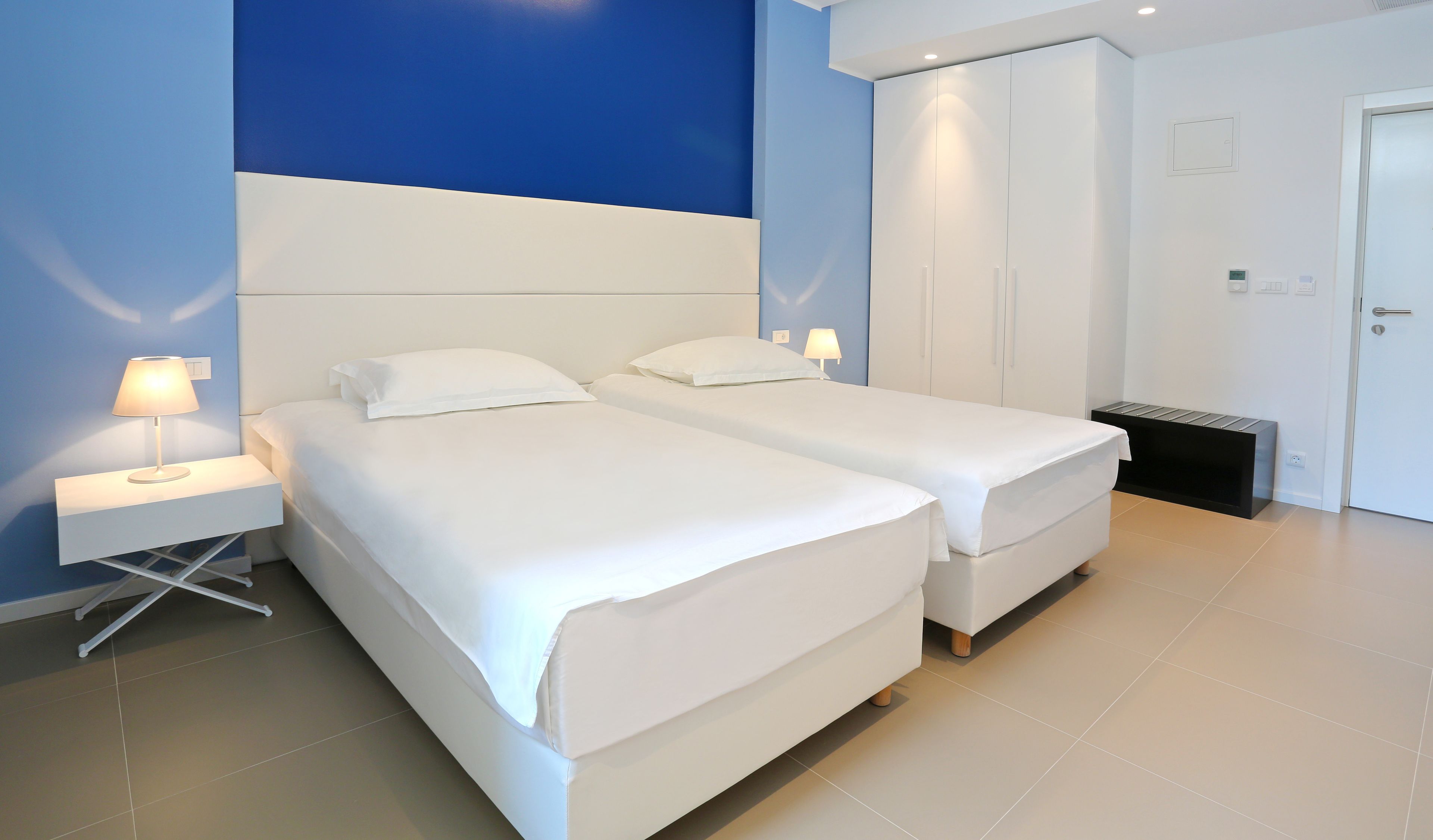 Une chambre d’hôtel avec deux lits, une table de chevet avec une lampe, une armoire et une poubelle.
