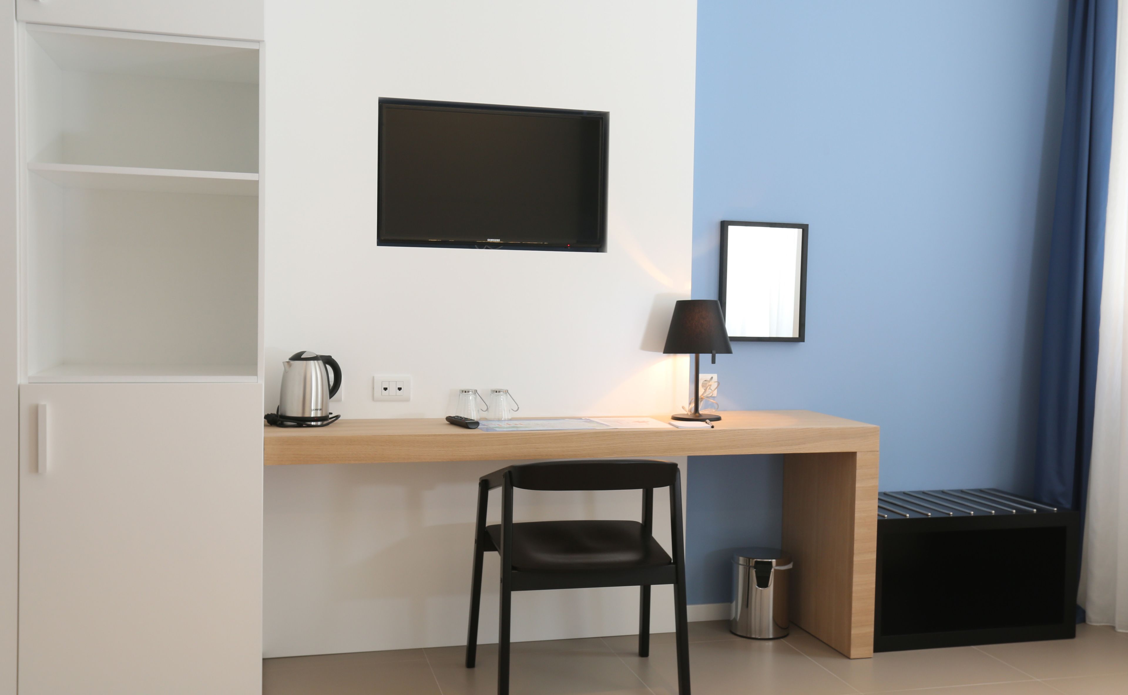 Una habitación de hotel moderna con un escritorio de madera, una silla negra, un televisor de pantalla plana en la pared, una mesita de noche con una lámpara y un cesto de basura.