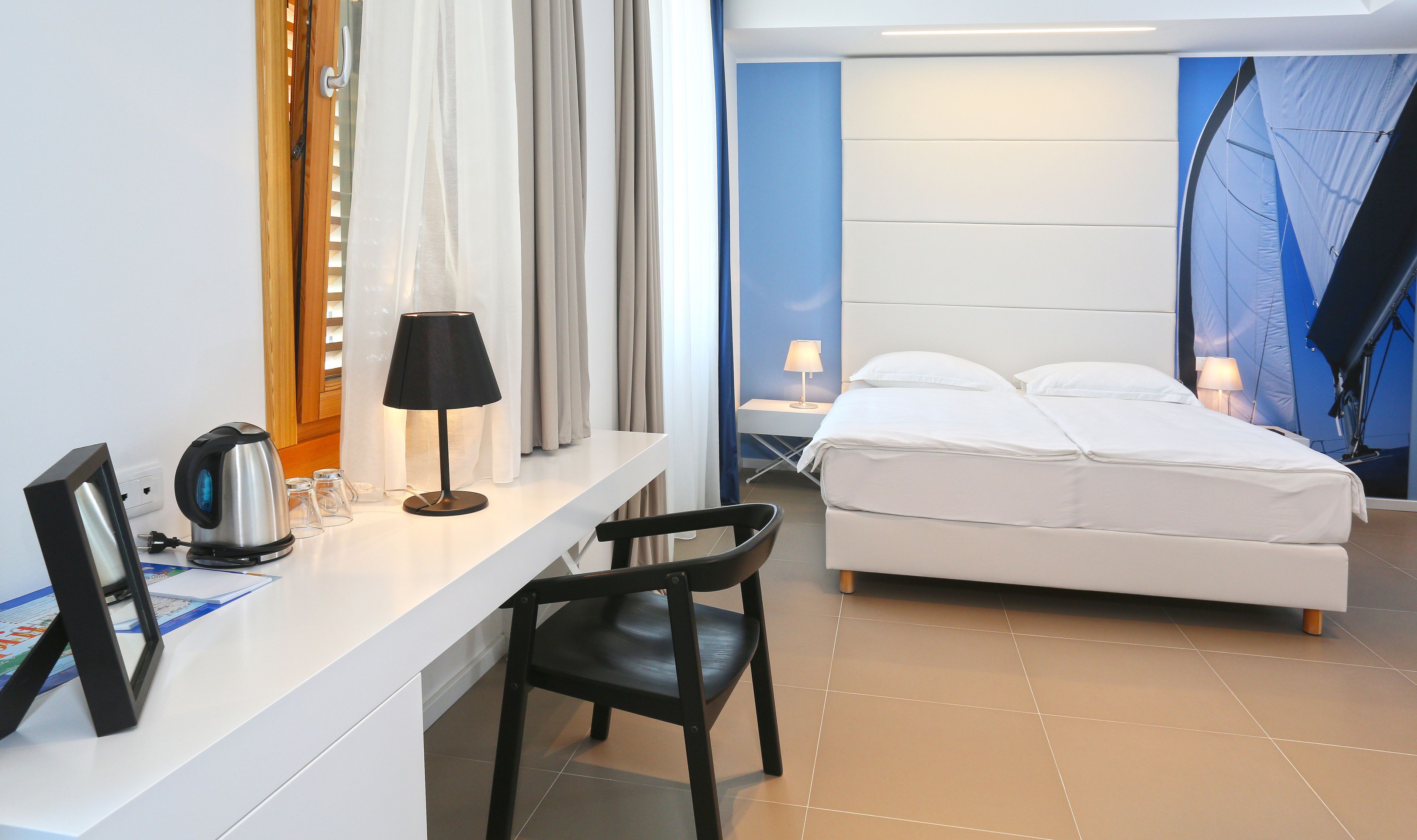 A hotel room with a bed, a desk, a chair, a kettle, and a lamp.