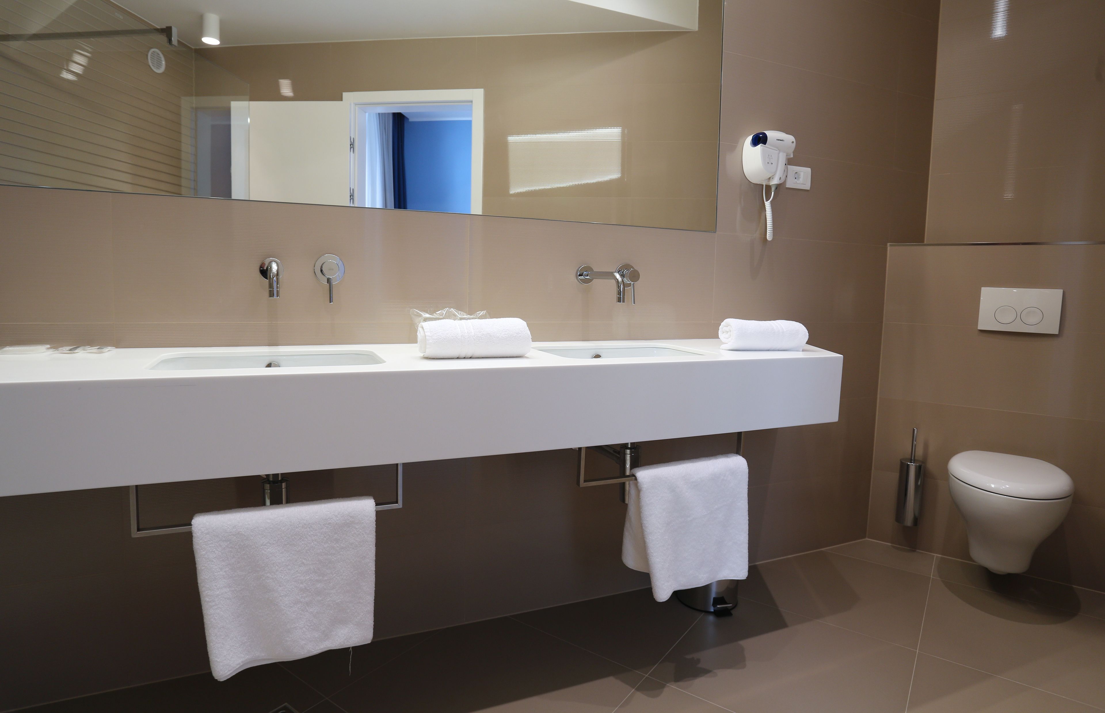 A modern bathroom with a large mirror, two sinks, a hairdryer, and a toilet.