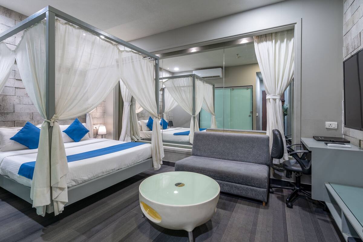 A hotel room with a four-poster bed, a grey sofa, a round table, and a desk with a chair.