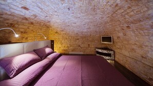 Un dormitorio con pared de piedra, una cama con ropa de cama púrpura, una mesita de noche y un televisor en la pared.