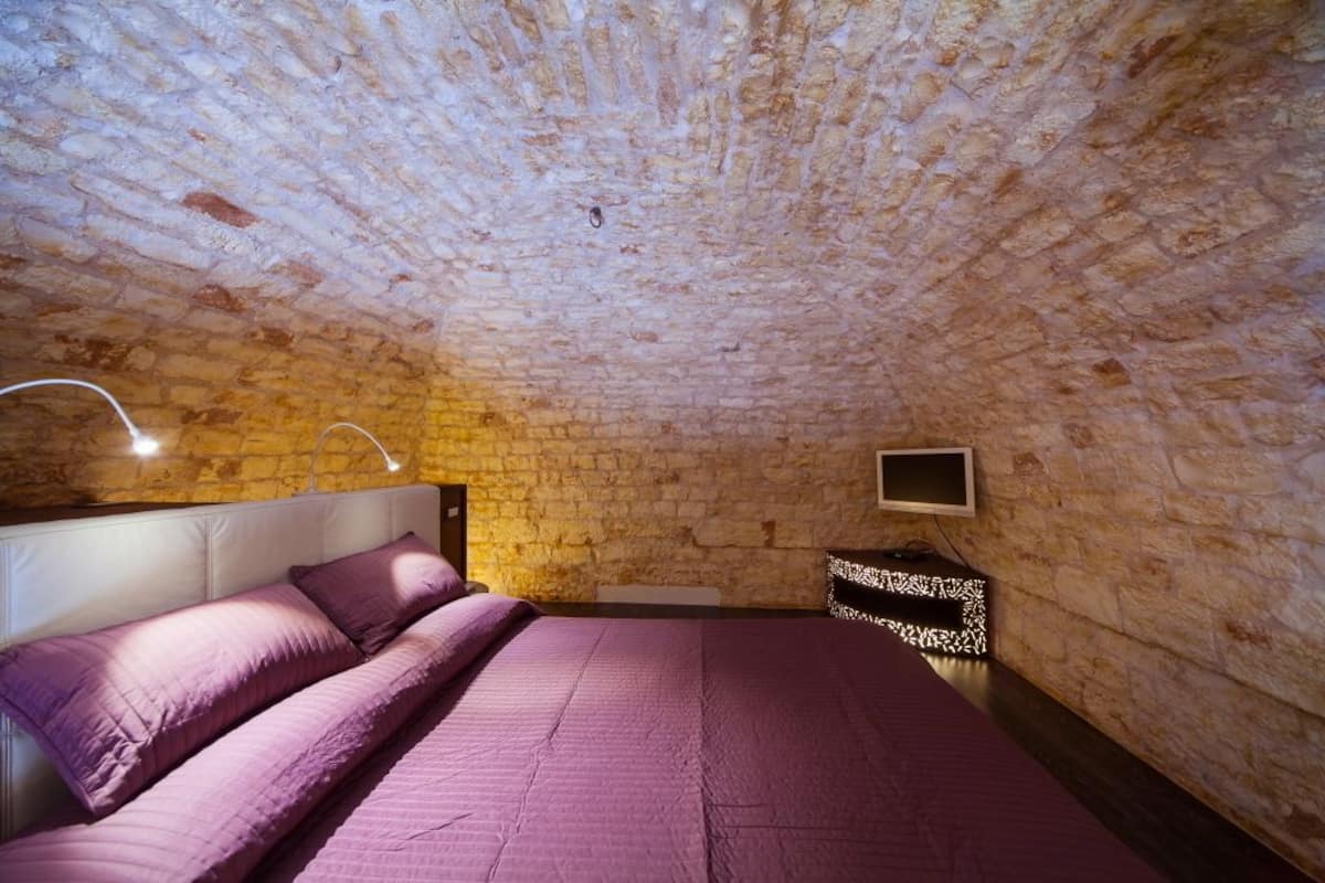 Un dormitorio con pared de piedra, una cama con ropa de cama púrpura, una mesita de noche y un televisor en la pared.