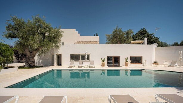 Una piscina con sillas de descanso, un edificio de fachada blanca y follaje circundante.