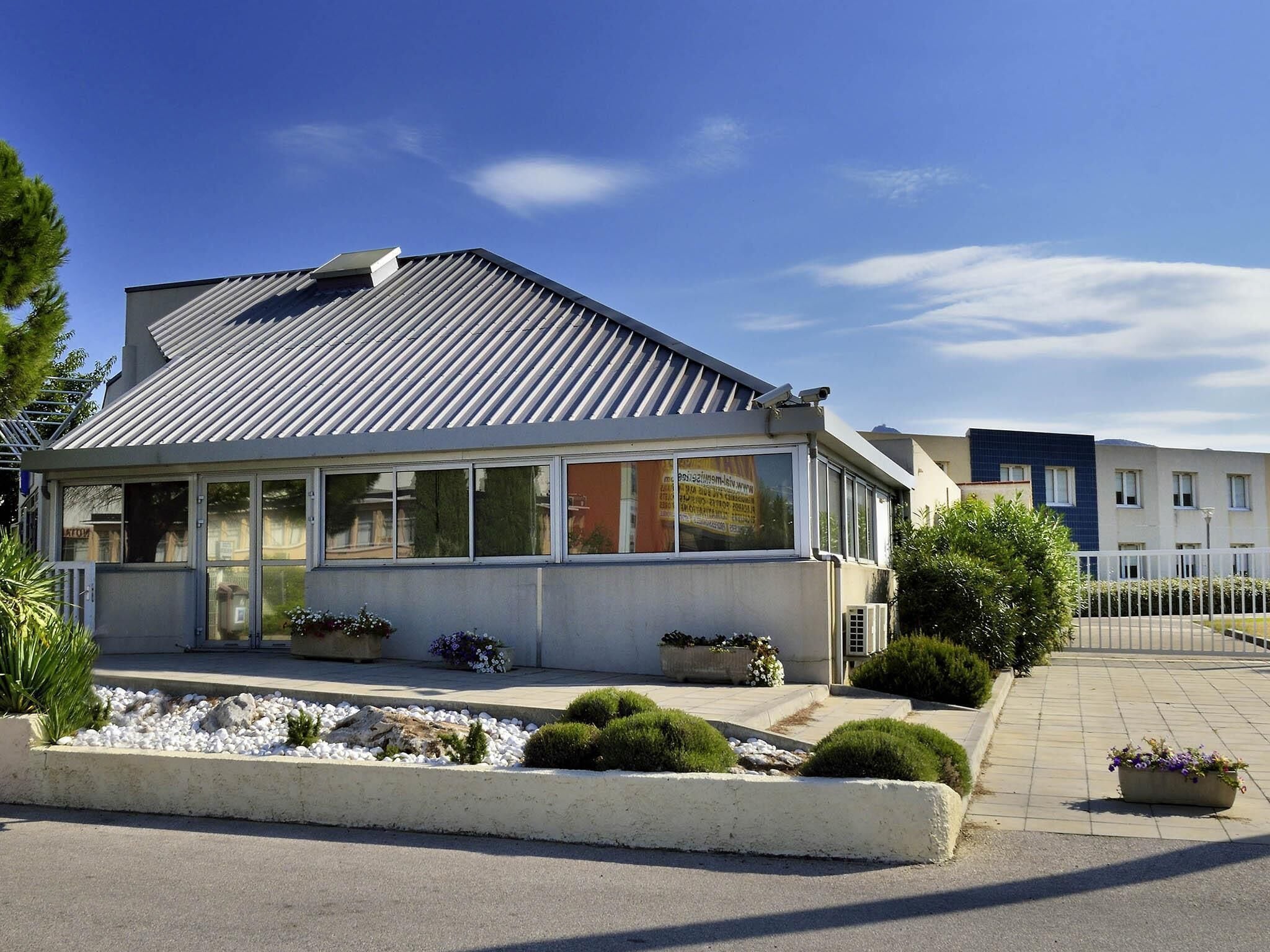 A modern building with a glass facade, a metal roof, and a paved entrance area with planters.