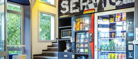 A vending machine with a beverage selection, a staircase, and a window with a view of greenery.
