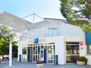 Entrada de hotel com fachada de vidro moderna, uma placa azul indicando ser um hotel econômico, e uma cobertura acima da entrada.