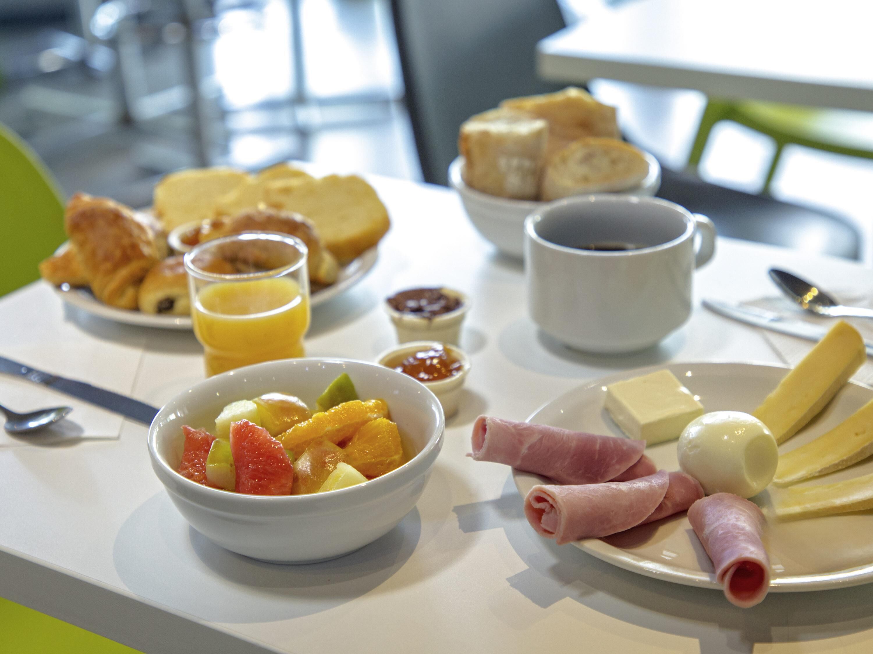 Una mesa de desayuno con diversos alimentos, incluyendo un tazón de fruta, rebanadas de queso, jamón y pan.
