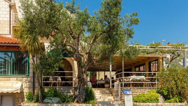 Un espace extérieur aménagé avec des murs en pierre, comprenant des tables et des chaises, un escalier menant à un niveau supérieur et un arbre au premier plan.
