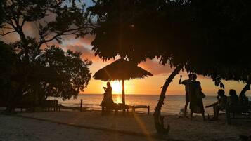 A beach scene with a sunset, a thatched umbrella, and people relaxing.