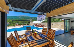 A modern outdoor pool area with wooden furniture and a table set for dining, overlooking a scenic mountainous landscape. - Cozy apartment in Kastel Stari (Kastel Stari)