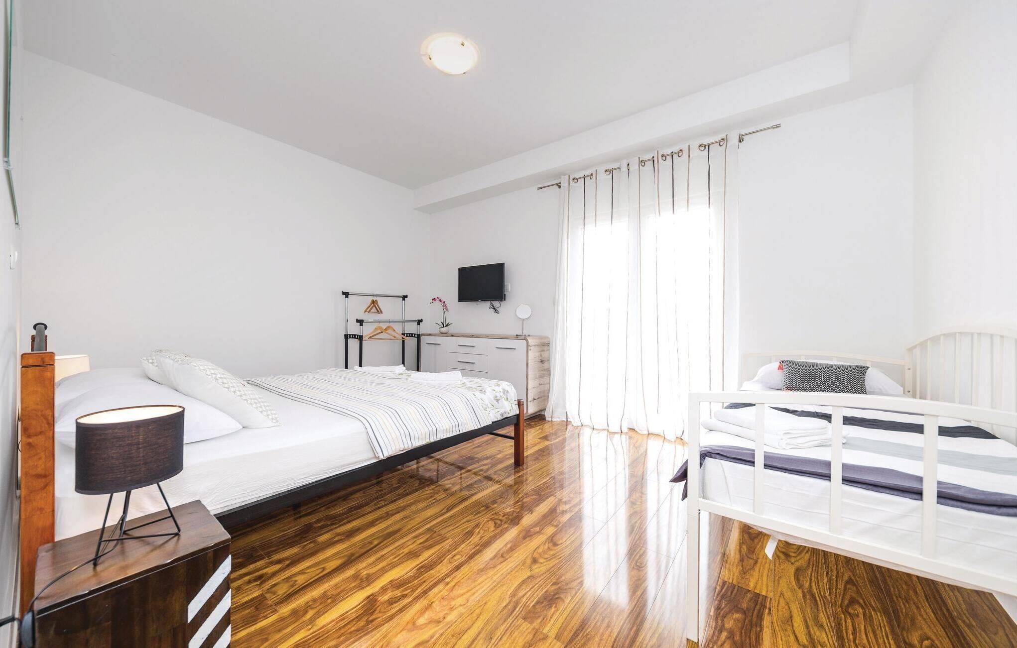 A bedroom with a wooden floor, a bed with a white mattress, a bedside table with a lamp, a dresser, and a TV on the wall.