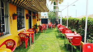 An outdoor seating area with red chairs and tables, a yellow building with a striped awning, and potted plants. - Hostal Miramar (Barbate)
