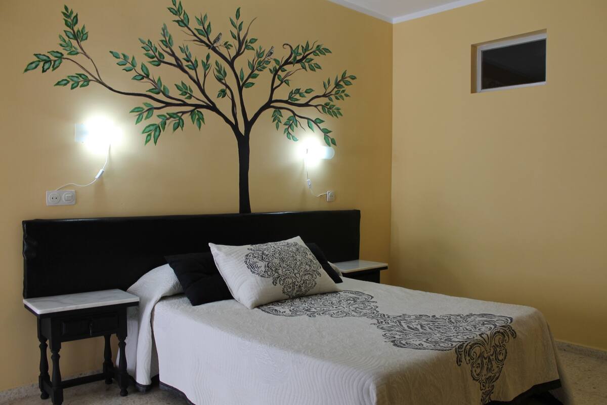 A bedroom with a bed, two bedside tables, a wall-mounted lamp, and a small window.