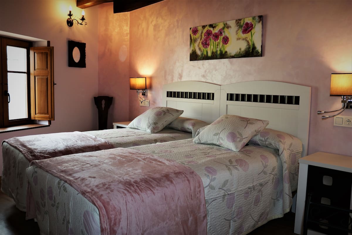 A bedroom with two beds, a painting of flowers, wall sconces, and a window with an open door.