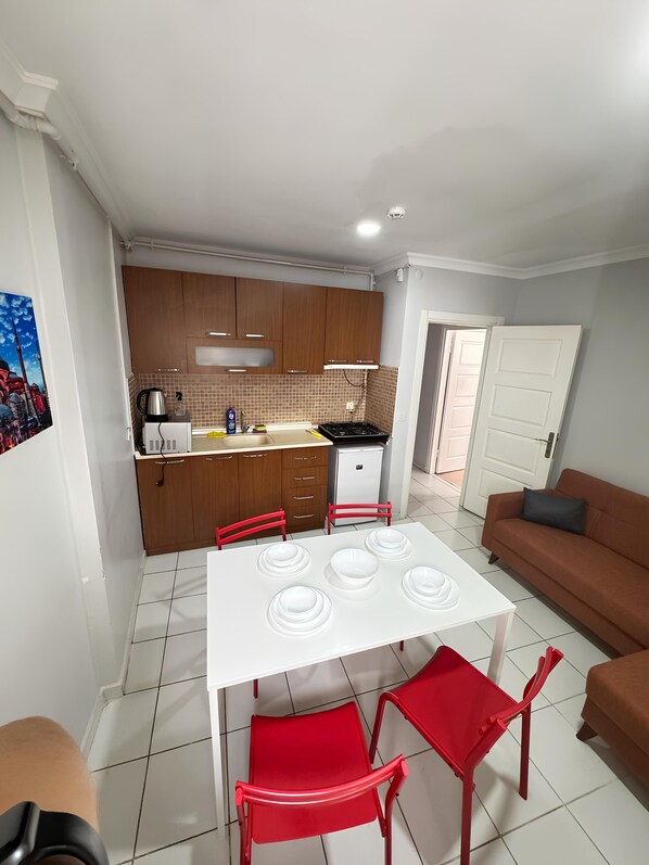 Un espacio habitable compacto con kitchenette, un área de comedor con mesa blanca y sillas rojas, y un sofá.