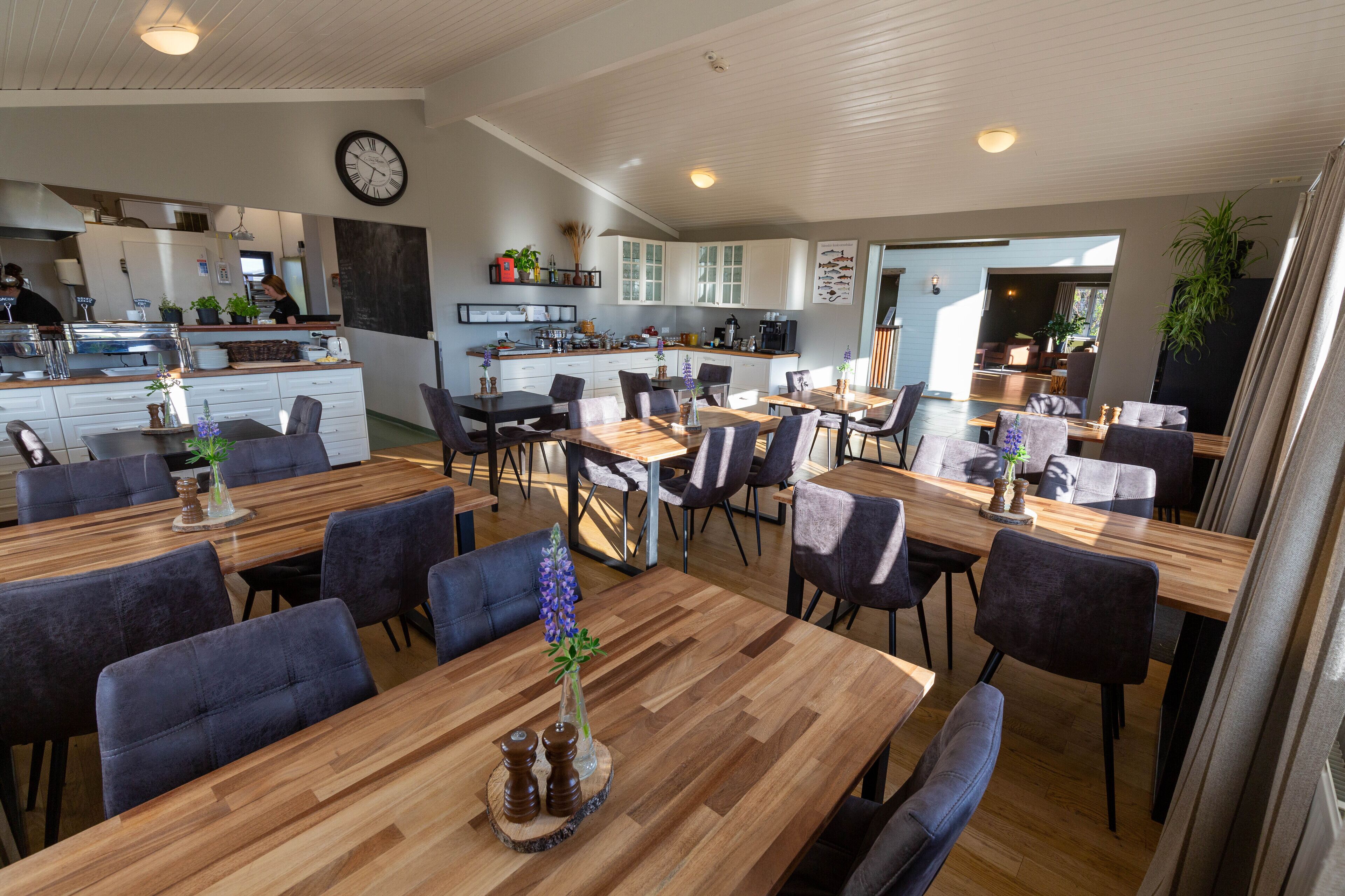 A spacious dining area with multiple wooden tables and dark upholstered chairs, a kitchen in the background, and a large clock on the wall.
