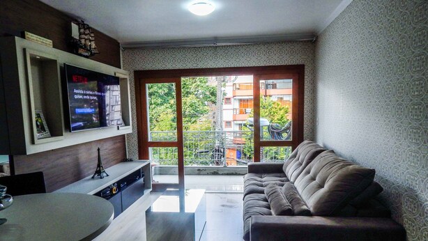 A modern living room with a sofa, a TV mounted on the wall, and a balcony with a view of buildings. - Charme Apartamento Centro de Gramado (Gramado)
