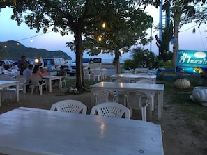 An outdoor dining area with tables and chairs, a tree, and a view of the sea.