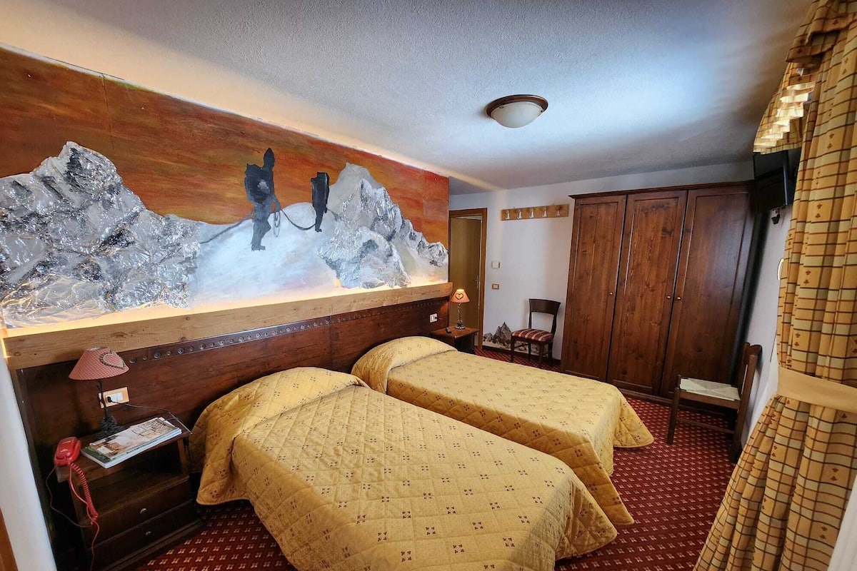 A hotel room with two beds, a wooden headboard, a painting of mountains, a wooden wardrobe, and a red patterned carpet.