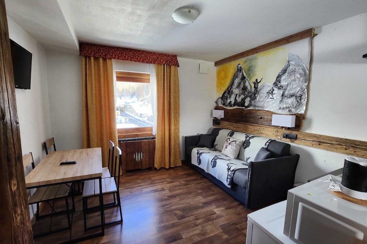 A room with a wooden dining table, a sofa with patterned cushions, a painting of mountains, and a white appliance.