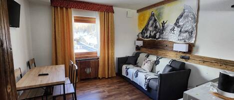 Una habitación con una mesa de comedor de madera, un sofá con cojines estampados, un cuadro de montañas y un electrodoméstico blanco.
