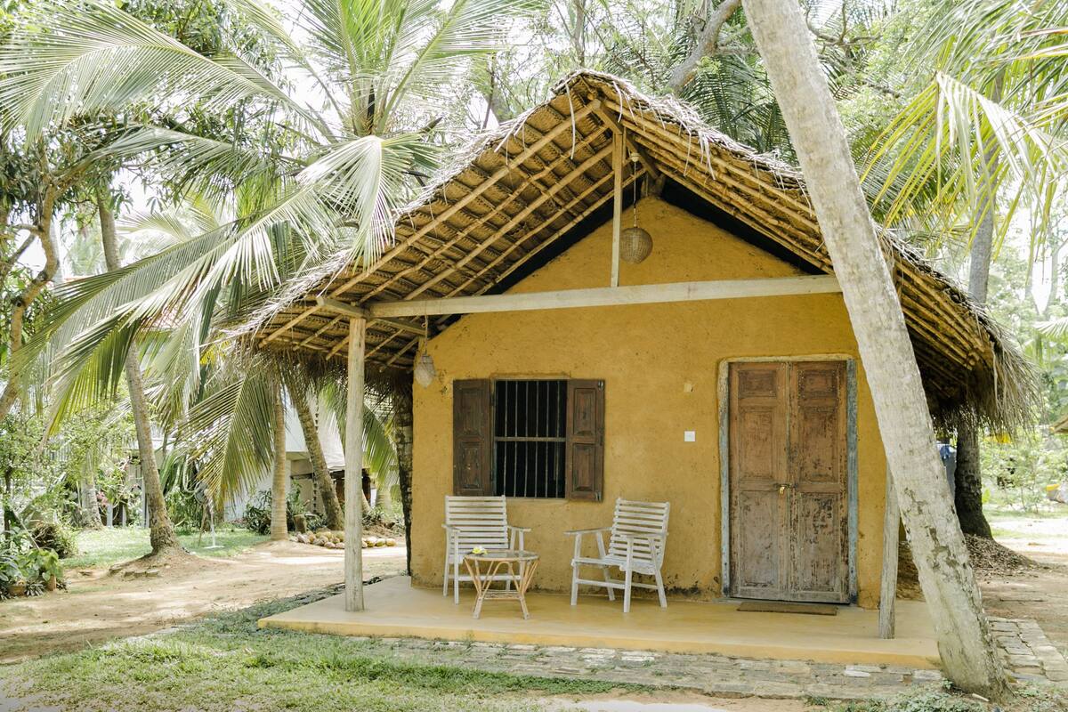 A small, single-story building with a thatched roof, two white chairs, and a wooden door.