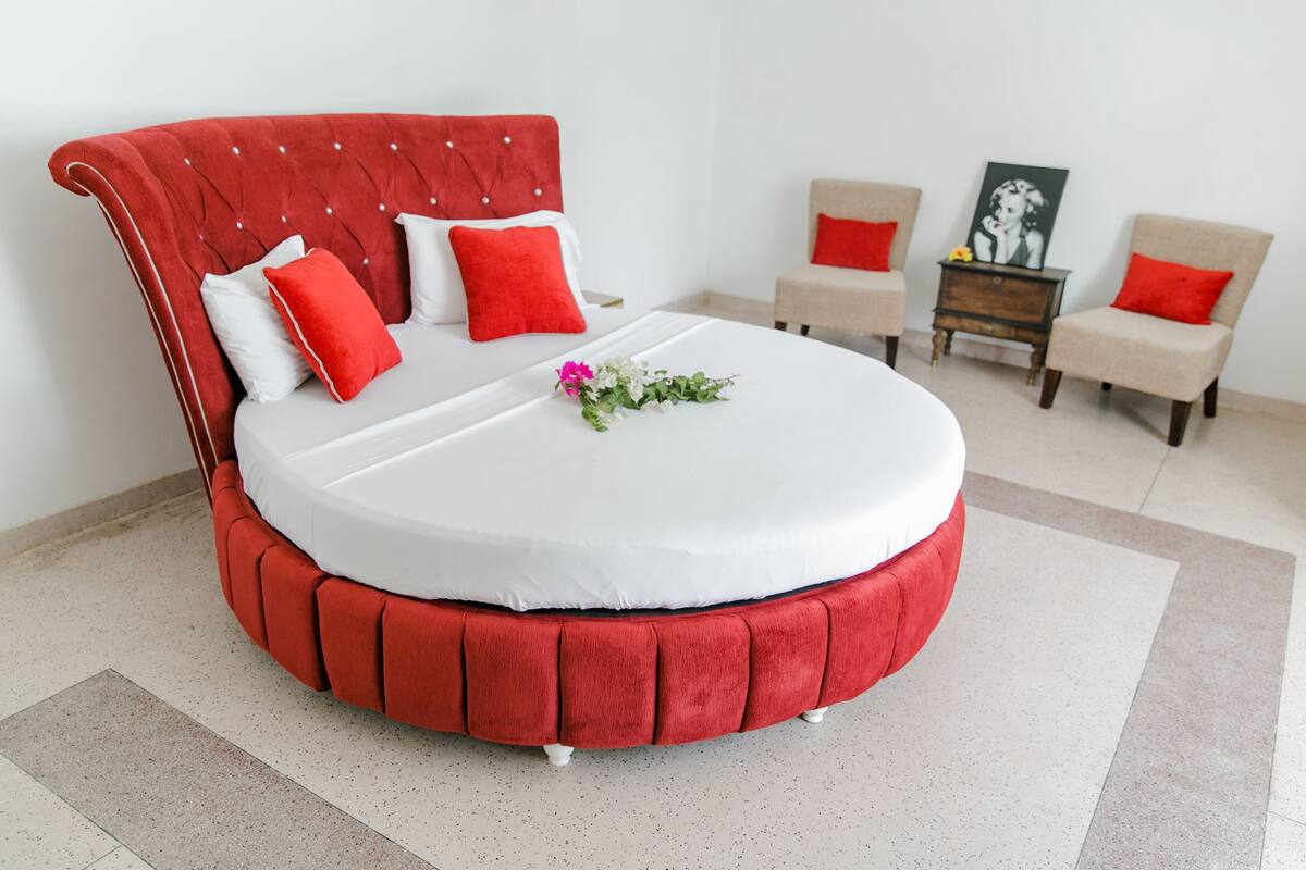 A round bed with a red upholstered headboard and white bedding, flanked by two beige chairs with red cushions, and a small wooden side table with a framed picture.