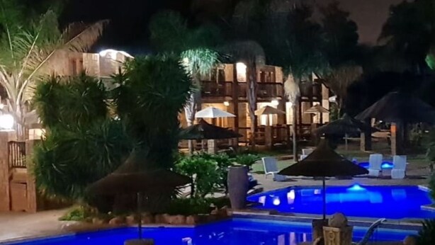 A nighttime pool area with lounge chairs and umbrellas, surrounded by palm trees and a building with multiple balconies. - Bushmans Rock Country Lodge (Roodeplaat)
