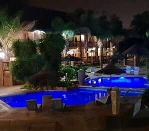 Zona de piscina nocturna con sillones de descanso y sombrillas, rodeada de palmeras y un edificio con varios balcones.